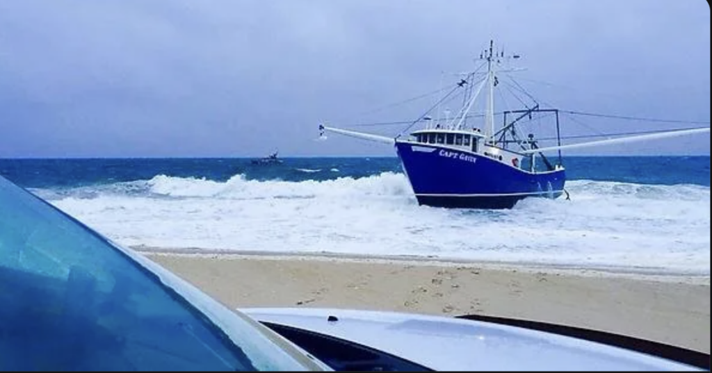 A blue and white fishing vessel named "CAPT GAVIN" is beached on a sandy shore with foamy waves. A smaller boat is visible in the background. The edge of a vehicle's windshield is in the foreground under an overcast sky.