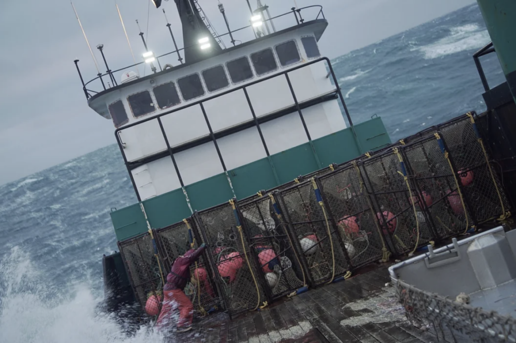 A fishing boat on rough seas with a person in red gear slipping on the wet deck. The deck is filled with crab pots and buoys. The sky is overcast, and waves are crashing against the vessel's side.