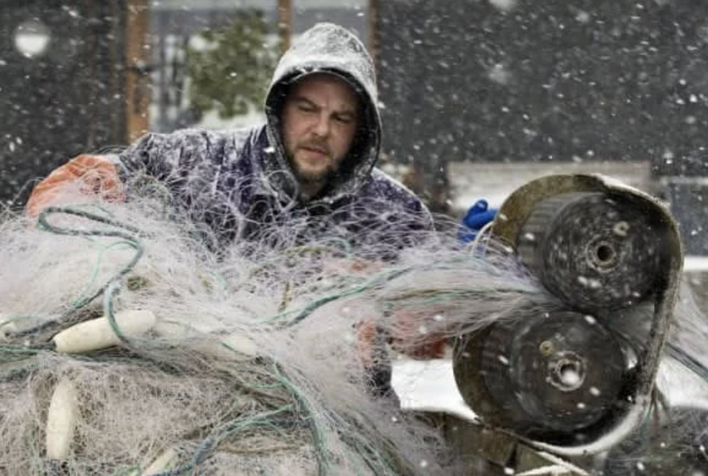 A person wearing a hooded jacket is untangling fishing nets in snowy weather. Snowflakes are falling around, and the person appears focused on the task. The background shows a dark building partially covered in snow.