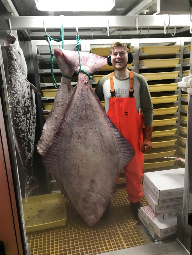 A person in orange overalls and a gray shirt stands smiling in a storage area with a large fish hanging beside them. The background shows trays and boxes stacked neatly on shelves.