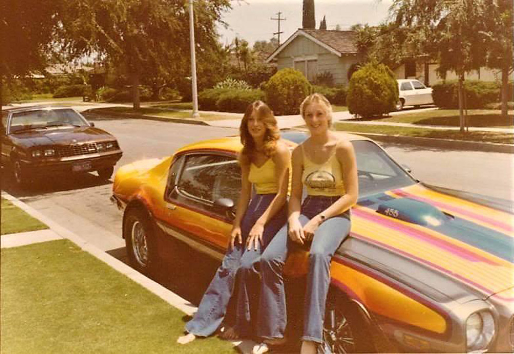 Two women in yellow tops and flared jeans sit on the hood of a colorful vintage car parked on a suburban street. Another car is parked nearby. Trees and houses line the background on a sunny day.