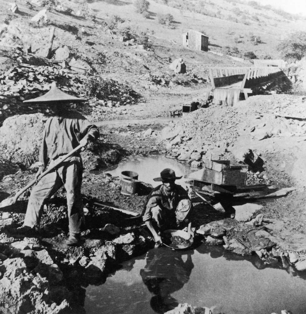 Two miners in a barren landscape, panning for gold near a small stream. One stands with a pan, the other leans over the water, both wearing hats. In the background, a wooden sluice box and a small shack can be seen.