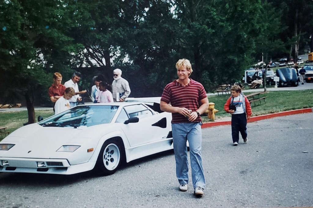 A man in a striped shirt walks in front of a white sports car, with several people in the background near tall trees. A child in a jacket follows him, and a picnic table and parked cars are visible nearby.