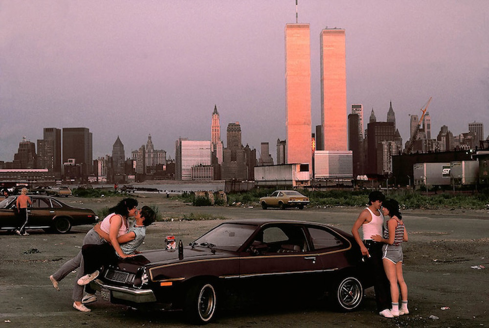 A 1970s scene with people gathered around a car against a backdrop of the New York City skyline, featuring the Twin Towers. A couple kisses on the hood, with others nearby, creating a nostalgic urban setting.