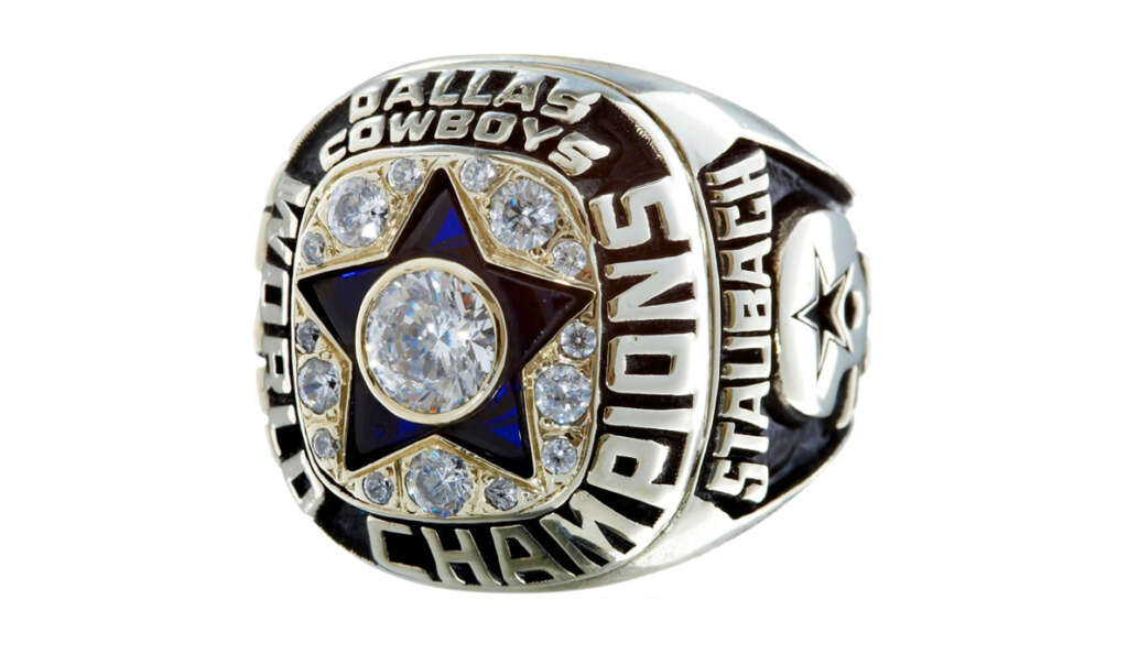 A silver championship ring featuring a star and diamond design at the center. The words "Dallas Cowboys World Champions" surround the star. "Staubach" is engraved on one side of the ring.