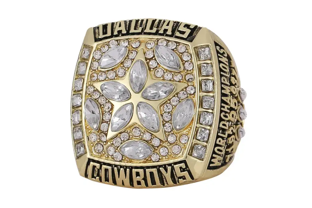 A gold Dallas Cowboys Super Bowl ring featuring a star design with large and small diamonds. The words "Dallas Cowboys" and "World Champions" are engraved in bold letters around the star.