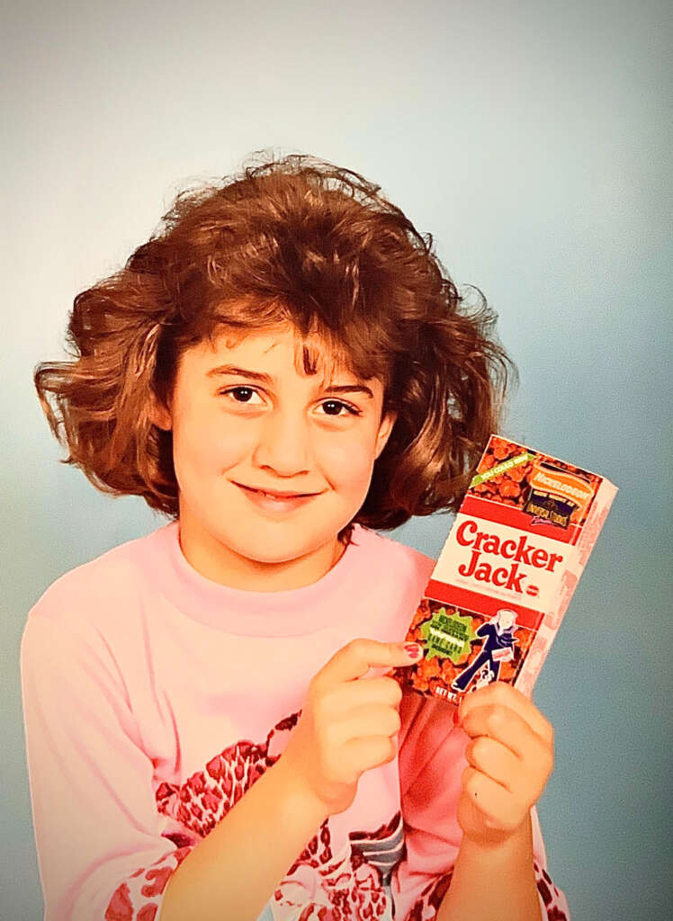 A child with curly hair smiles while holding a Cracker Jack box. They are wearing a pink shirt with a large red design. The background is plain light blue.