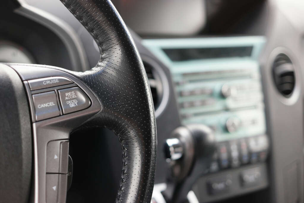 Close-up of a car's steering wheel with buttons for cruise control on the left. In the background, the car's dashboard is visible, featuring a radio with dials and knobs. The focus is mainly on the steering wheel.