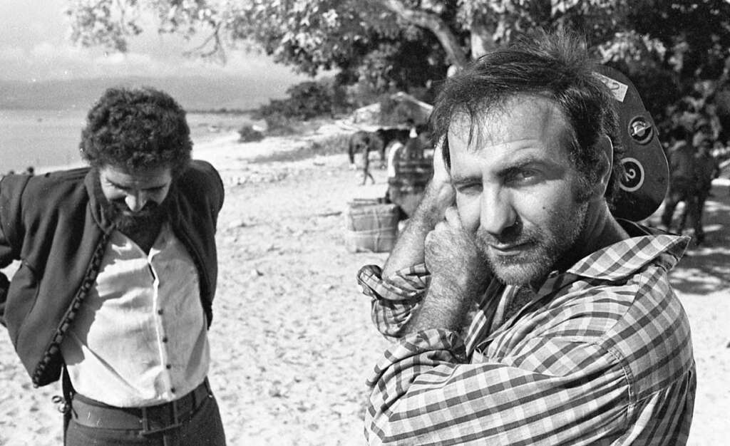 Two men stand on a beach. The man in the foreground holds a film camera on his shoulder and wears a checkered shirt, looking toward the camera. The other man, wearing a jacket, looks down. Horses and trees are in the background.