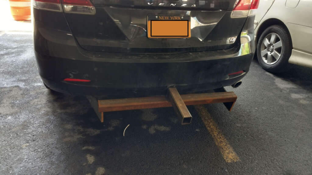 A black car with a customized, rusted metal hitch protruding from the rear bumper in a parking lot. The license plate is partly covered. Another vehicle is parked beside it.