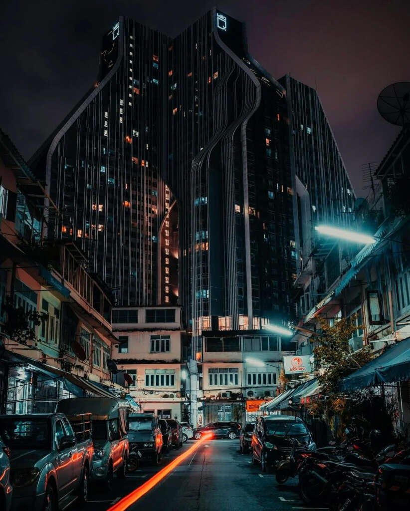 A tall, uniquely designed skyscraper with glowing windows towers over a busy, narrow street at night. Vehicles and motorbikes line the street, with tail lights creating a red streak on the road. Old-style buildings flank the street.