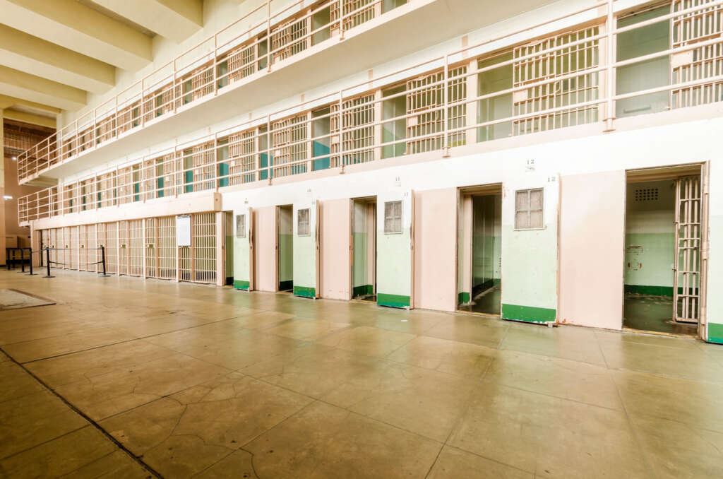 Empty prison hallway with a row of small cells, each with a barred door. The walls are pale, and there are two levels with a walkway above. The setting is well-lit and spacious, reflecting an older style of prison architecture.