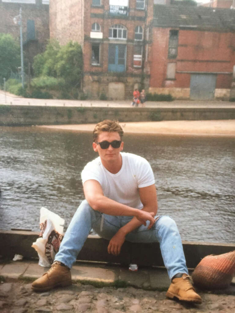 Man sitting on a riverside cobblestone path, wearing sunglasses, a white t-shirt, jeans, and boots. He sits casually with a brick wall and old buildings in the background. A bag is next to him, and the river flows behind.