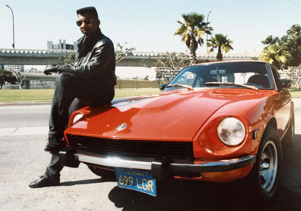 A person wearing a black leather jacket, gloves, and sunglasses sits on the hood of a red vintage sports car with a blue license plate. Palm trees and a bridge are visible in the background.