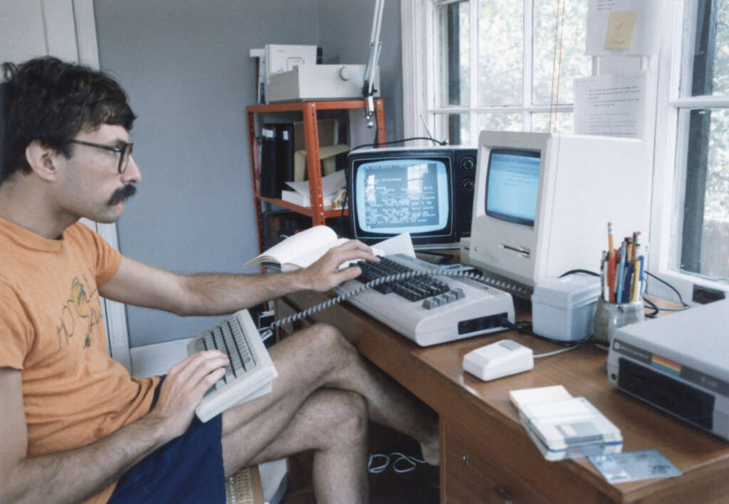 A person with glasses and a mustache is using a vintage computer setup. They're seated at a wooden desk with two monitors, a keyboard, mouse, floppy disks, and office supplies. A window is in the background, and a bookshelf is nearby.