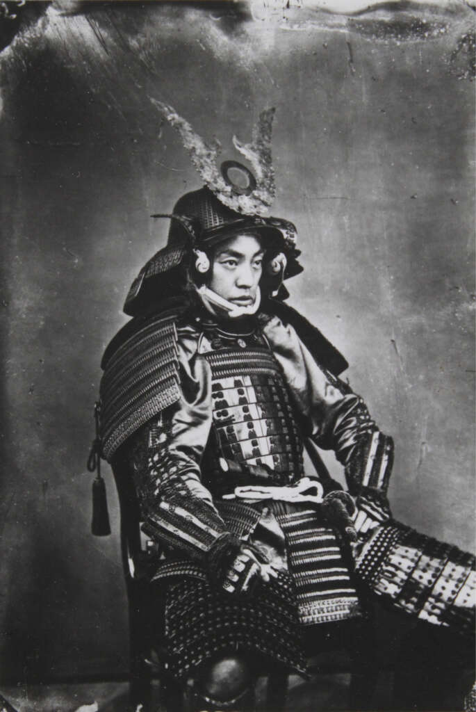 A black and white historical photograph of a man in full samurai armor, featuring a detailed helmet with a decorative crest. He is seated, looking to the side with a serious expression, in front of a plain backdrop.