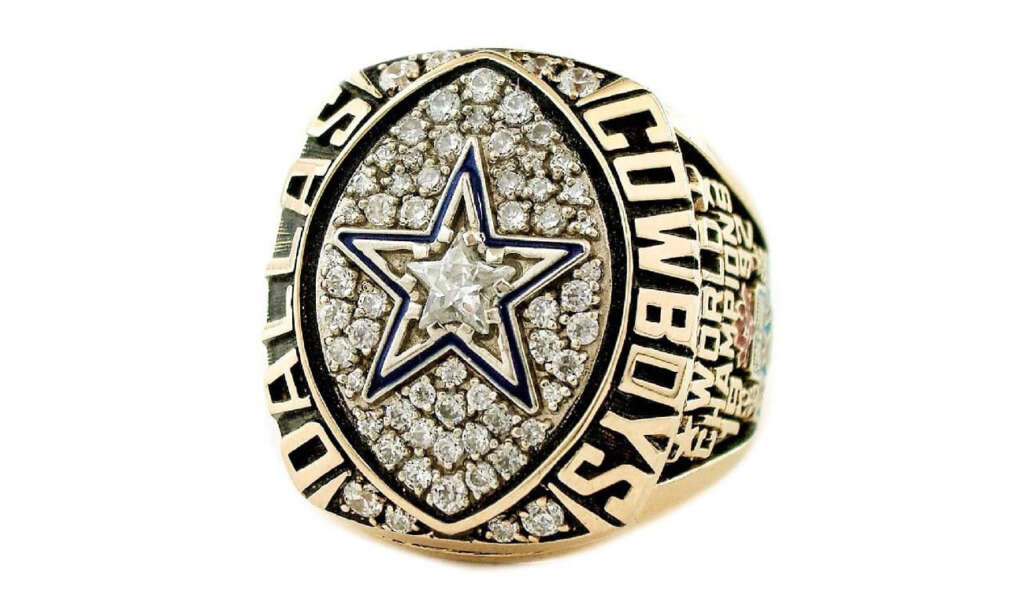 Gold Dallas Cowboys championship ring with a large star emblem in the center, surrounded by diamonds. The words "DALLAS COWBOYS" are inscribed around the star. The ring has intricate detailing on the sides.