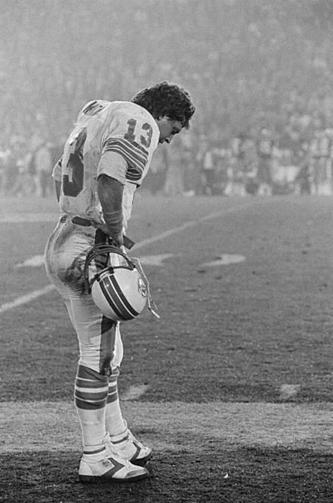 Football player in a number 13 uniform stands on the field, holding his helmet. The background shows a blurred crowd, and the setting seems to be a stadium with evening lighting.