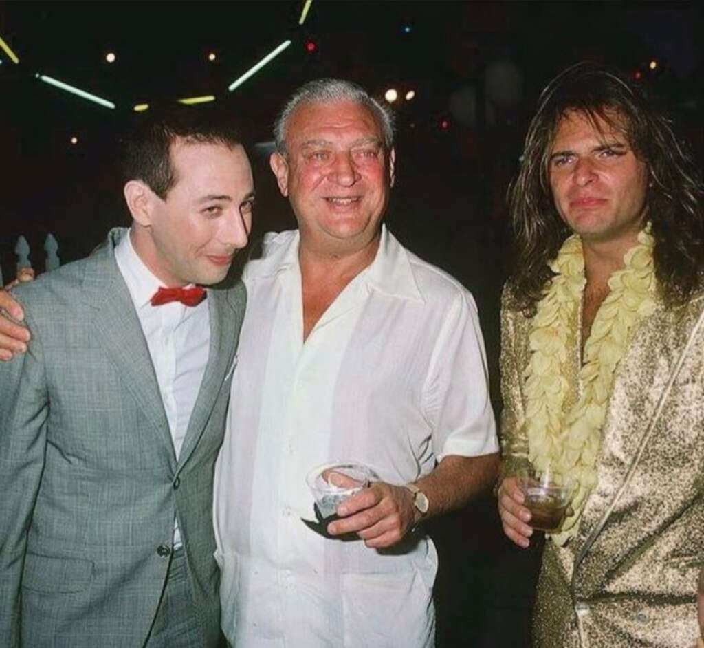 Three men are standing together at an event. The man on the left is in a grey suit with a red bow tie, the man in the middle is in a white shirt holding a drink, and the man on the right is wearing a gold outfit and a flower lei, holding a drink.