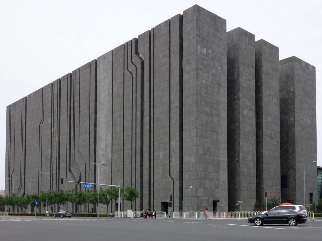 A large, modern, gray building with a geometric, block-like design and vertical lines running down its facade. Located at an intersection with a few vehicles and bicycles. Trees line the street in front of the building.