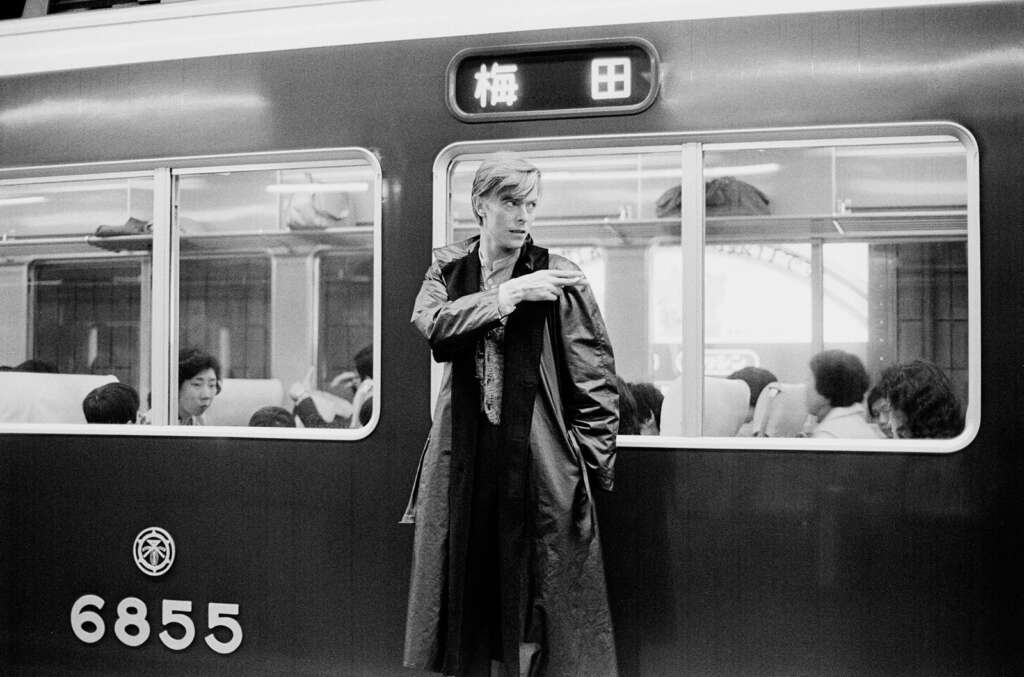 A person in a long coat stands leaning against the side of a train numbered 6855. They are looking to their left. Inside the train, passengers are seated, some looking out the window. The station name “梅田” (Umeda) is visible through the window.