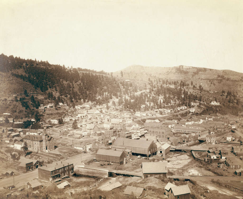 A sepia-toned aerial view of a small 19th-century town surrounded by hills and dense forests. The town features wooden buildings, dirt roads, and scattered structures, capturing a historical rural setting amidst the natural landscape.