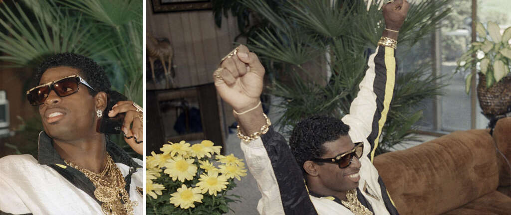 Deion Sanders wearing sunglasses and gold jewelry is sitting on a couch, talking on a phone. He is dressed in a white and black outfit with one arm raised joyfully. Yellow flowers and green plants are visible in the background.