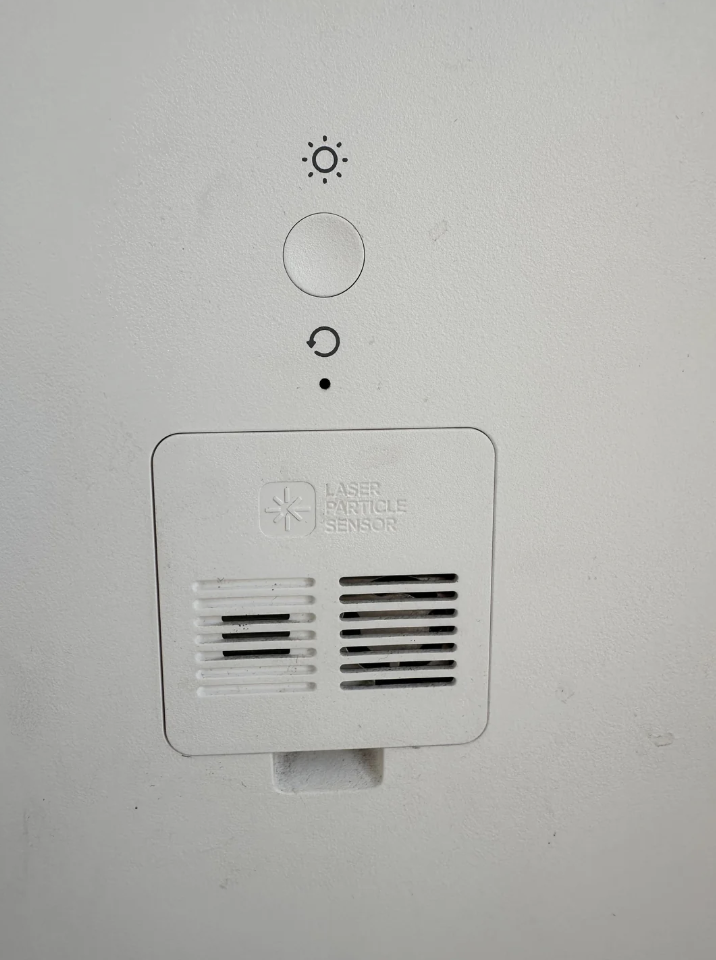 Close-up of a white wall-mounted device with a labeled panel "Laser Particle Sensor." It features two vent-like openings and indicator icons above, including a sun symbol and a circular arrow.