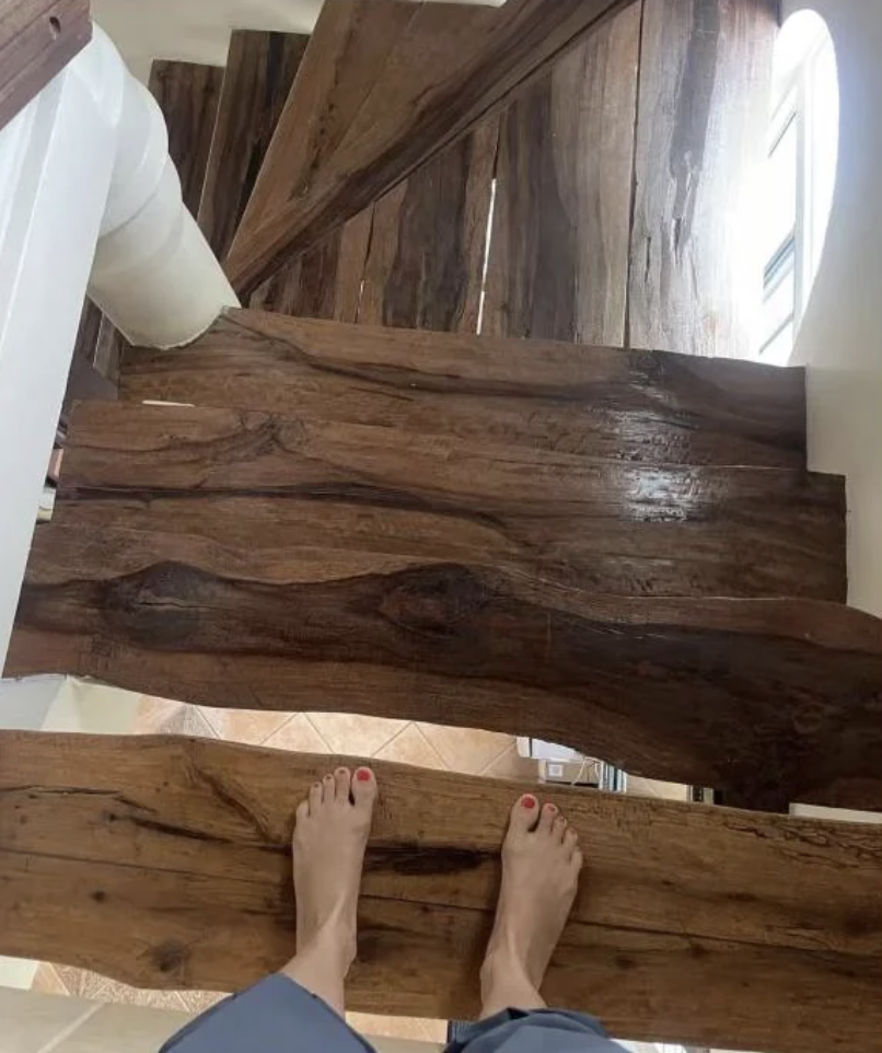 A person standing barefoot at the top of a narrow wooden staircase with a rustic finish. The stairs descend in a winding pattern against white walls and a window allows natural light to illuminate the space.