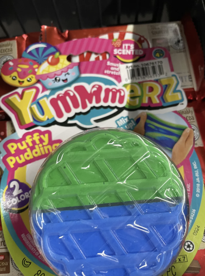 A package of Yummy Meerz Puffy Pudding is shown, featuring a blue and green squishy toy on top. The packaging design includes colorful text and cute graphics. It mentions that the toy is scented and contains two colors.