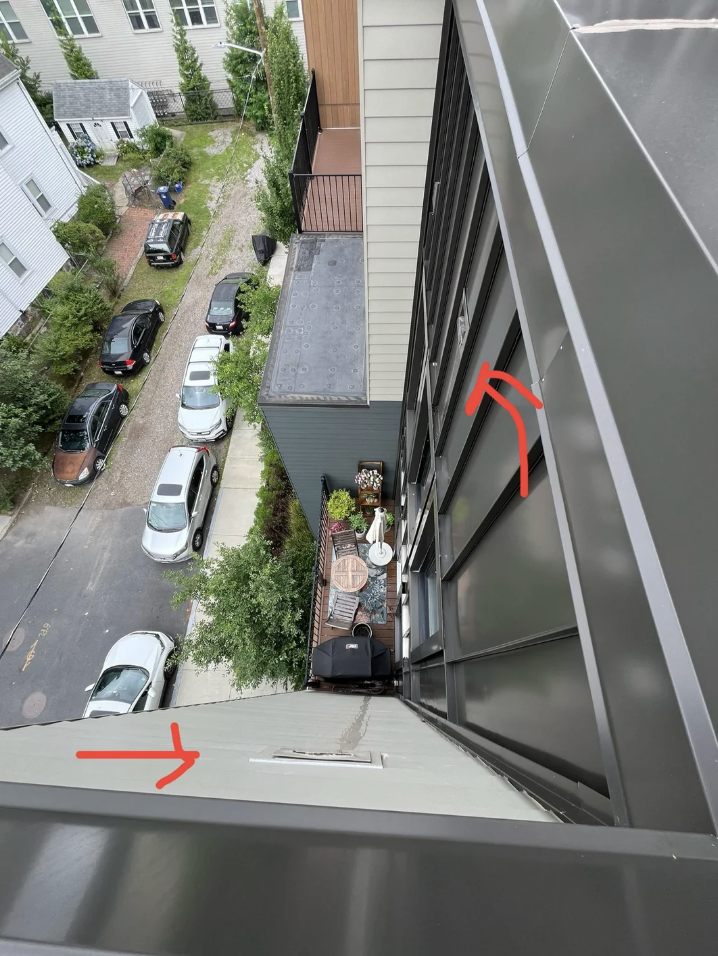 View from a high building showing a narrow alley with parked cars, balconies, and red arrows pointing to windows. A patio with a table and chairs is visible on one balcony.