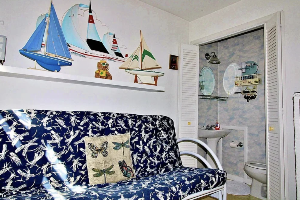 A room features a blue and white patterned sofa with a butterfly cushion. Above, a shelf displays model sailboats and a teddy bear. A small, open door leads to a bathroom with a sink and mirrored cabinet. Walls are white and light blue.