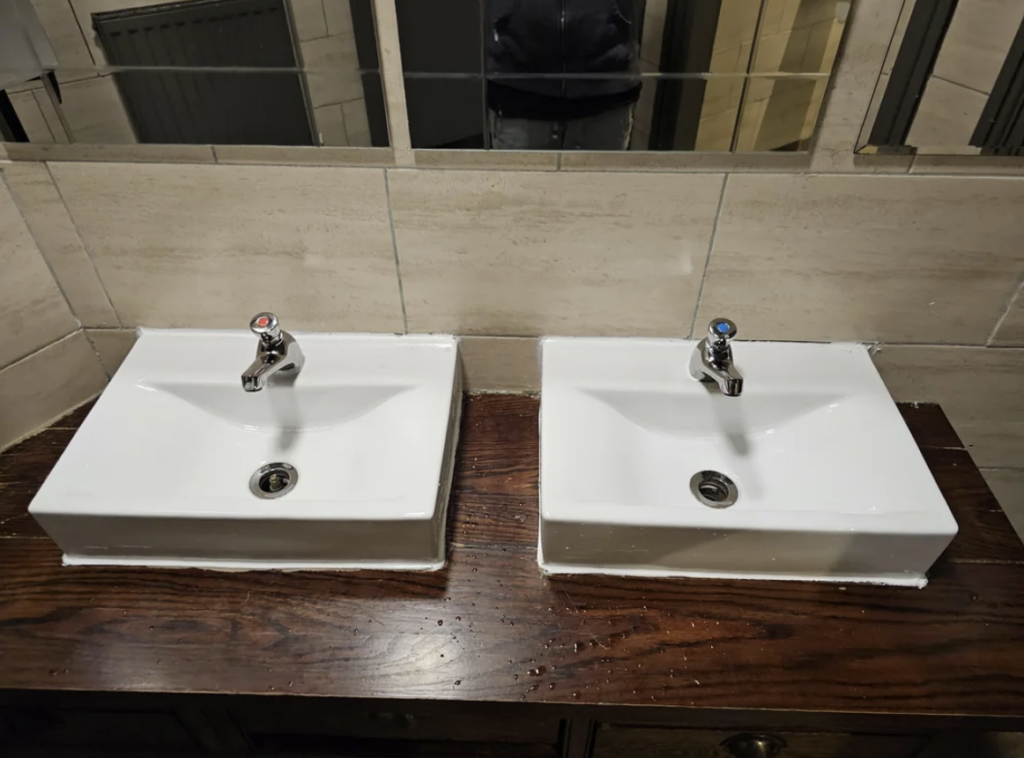 Two white rectangular sinks with single faucets sit side by side on a dark wooden countertop. Each faucet has a red and blue indicator for hot and cold water. The background features beige tiled walls and a partial view of a mirror.