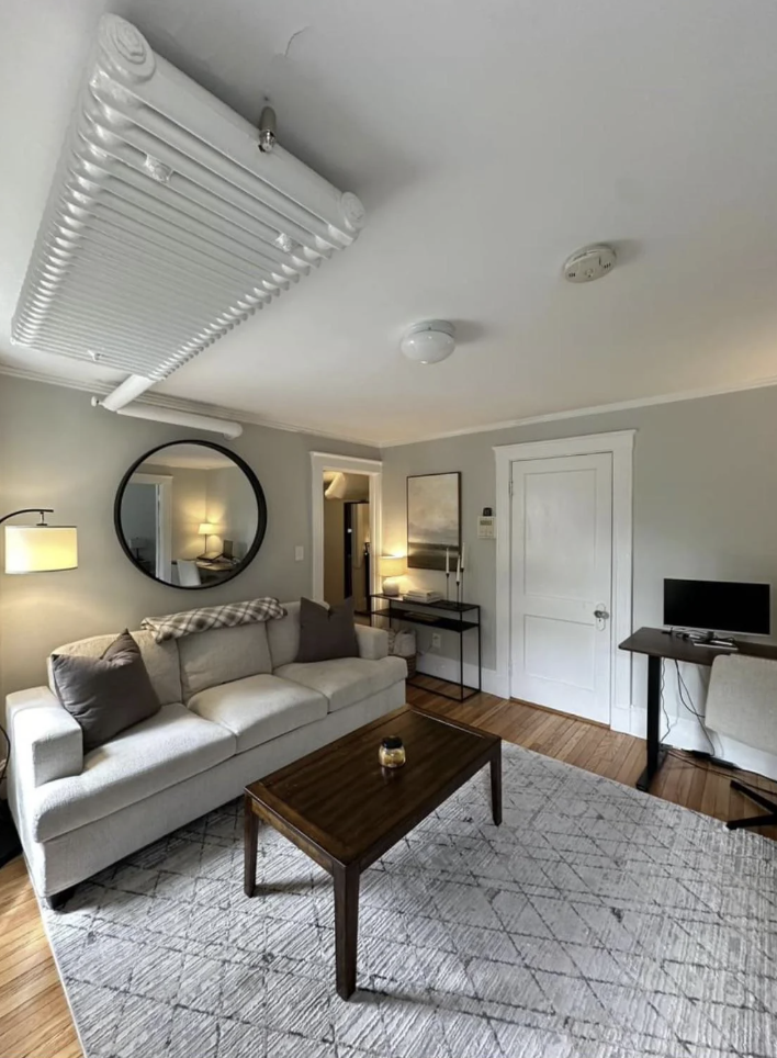 A cozy living room with a white sofa, a wooden coffee table, and a patterned rug. A round mirror hangs above the sofa. A small desk and computer are in the corner. The room has a modern light fixture and a neutral color palette.