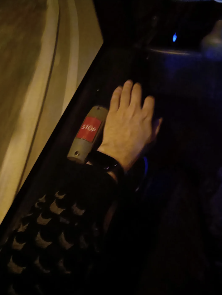 A hand reaches up to press a red "STOP" button on a vehicle wall, with dim lighting suggesting nighttime. The person wears a dark patterned sleeve and a black wristband. Lines of light streak outside the window, indicating movement.