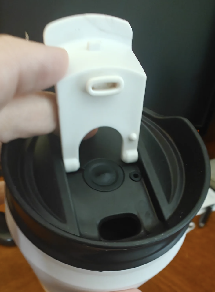 A hand is holding a white travel mug lid with a black interior. The lid is open, revealing the drinking spout. The background is a blurred dark surface, possibly a desk or table.