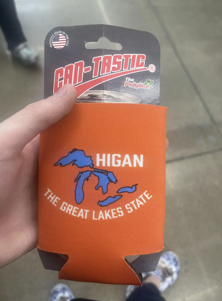 A hand holding an orange can cooler. It has a blue outline of the Great Lakes and the text "HIGAN: THE GREAT LAKES STATE." The packaging displays the brand "CAN-TASTIC" with an American flag emblem.