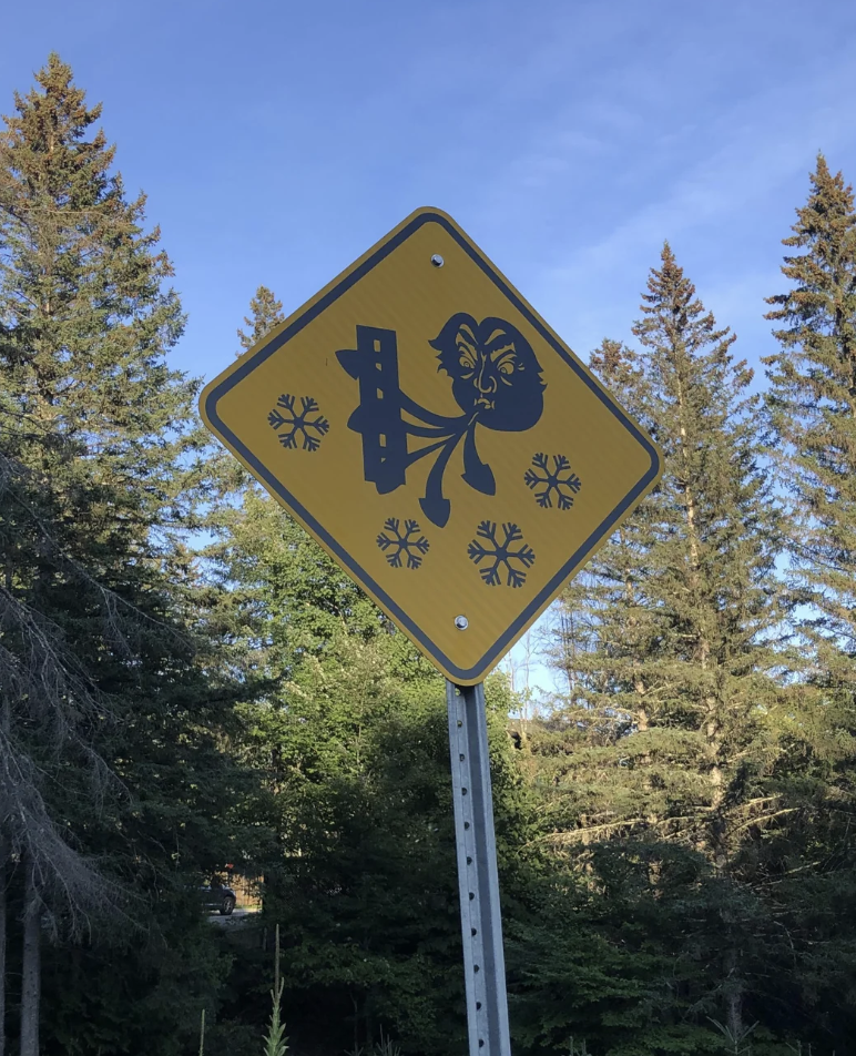 A yellow diamond-shaped sign depicts a human figure being blown by strong wind with snowflakes around. The sign is set against a background of tall evergreen trees and a clear blue sky.