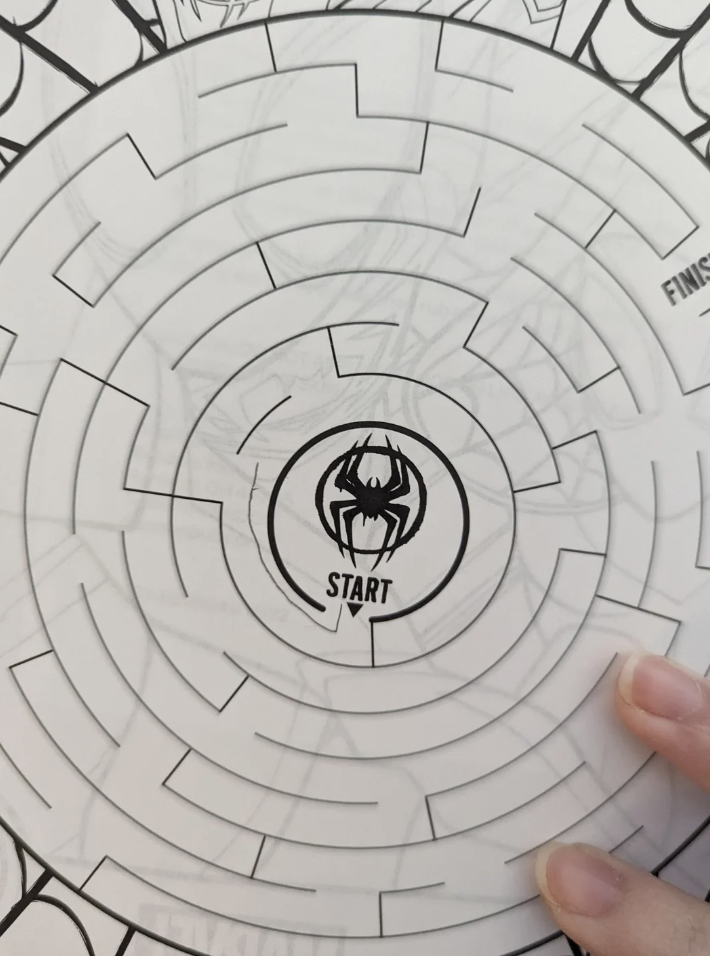 A hand holds a circular maze puzzle. The center of the maze is marked with "START" and a spider symbol. The maze path winds outward. The outer edge shows "FINISH" with a partial view of a character drawing underneath.