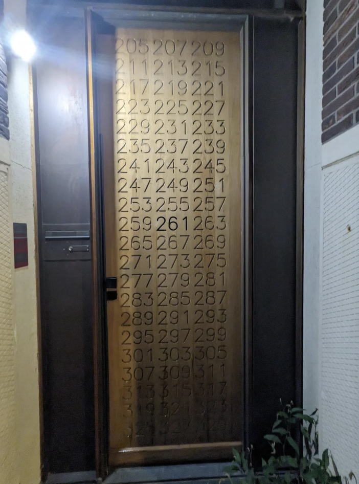 A door panel is covered with numbers arranged in a grid, from 205 to 319, with 261 highlighted at the center. The door is framed by a brick wall on the right and a light source above. Some plants are partially visible at the bottom.