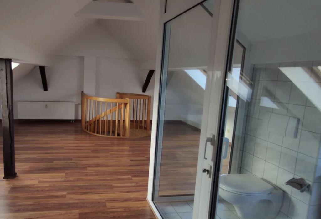 An attic room with hardwood floors, featuring a spiral wooden staircase, a large window, and a small bathroom with glass doors. The ceiling has exposed beams and slanted angles, creating a cozy atmosphere.