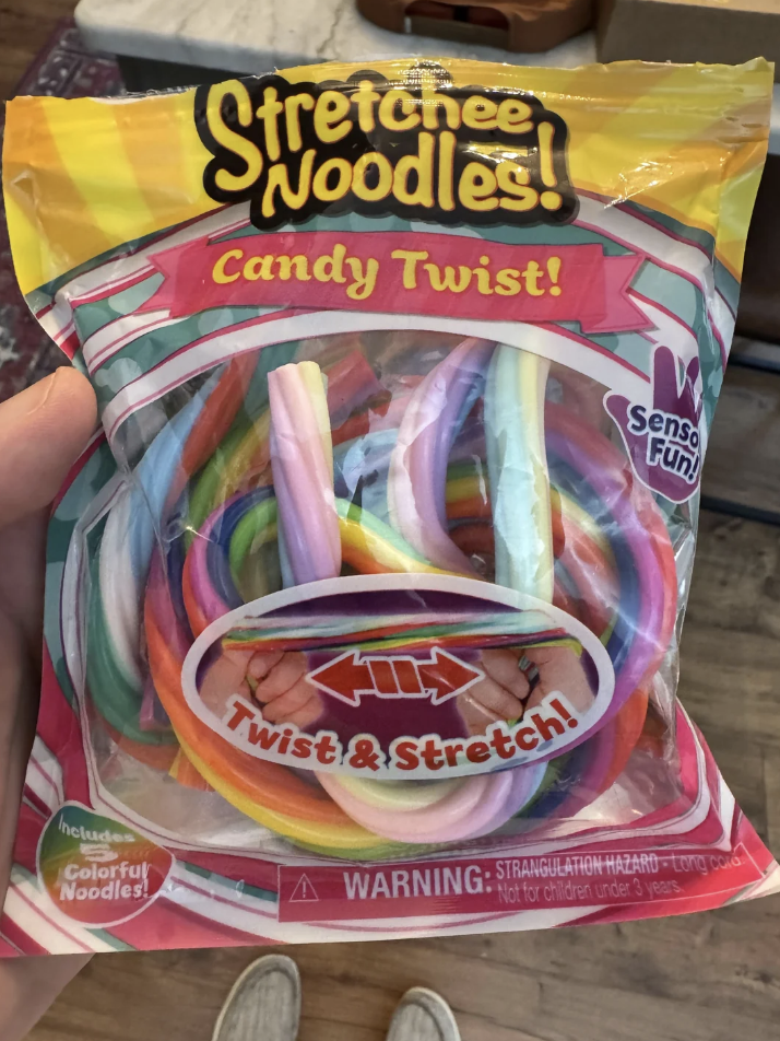 A package of "Stretchee Noodles! Candy Twist," featuring colorful, tubular candy pieces. The packaging highlights features like "Twist & Stretch" and includes a warning about a potential strangulation hazard.