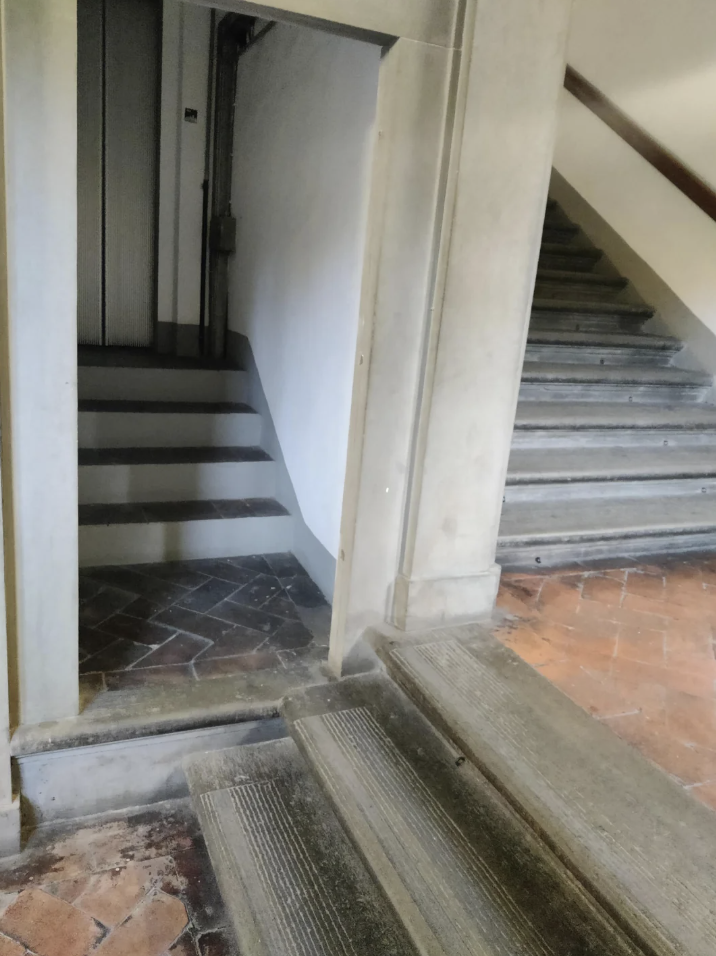 A narrow stairwell with brick and stone steps leading upward. Adjacent is a small elevator with its door open, positioned slightly lower than the first step. The walls are plain and the overall lighting is dim.