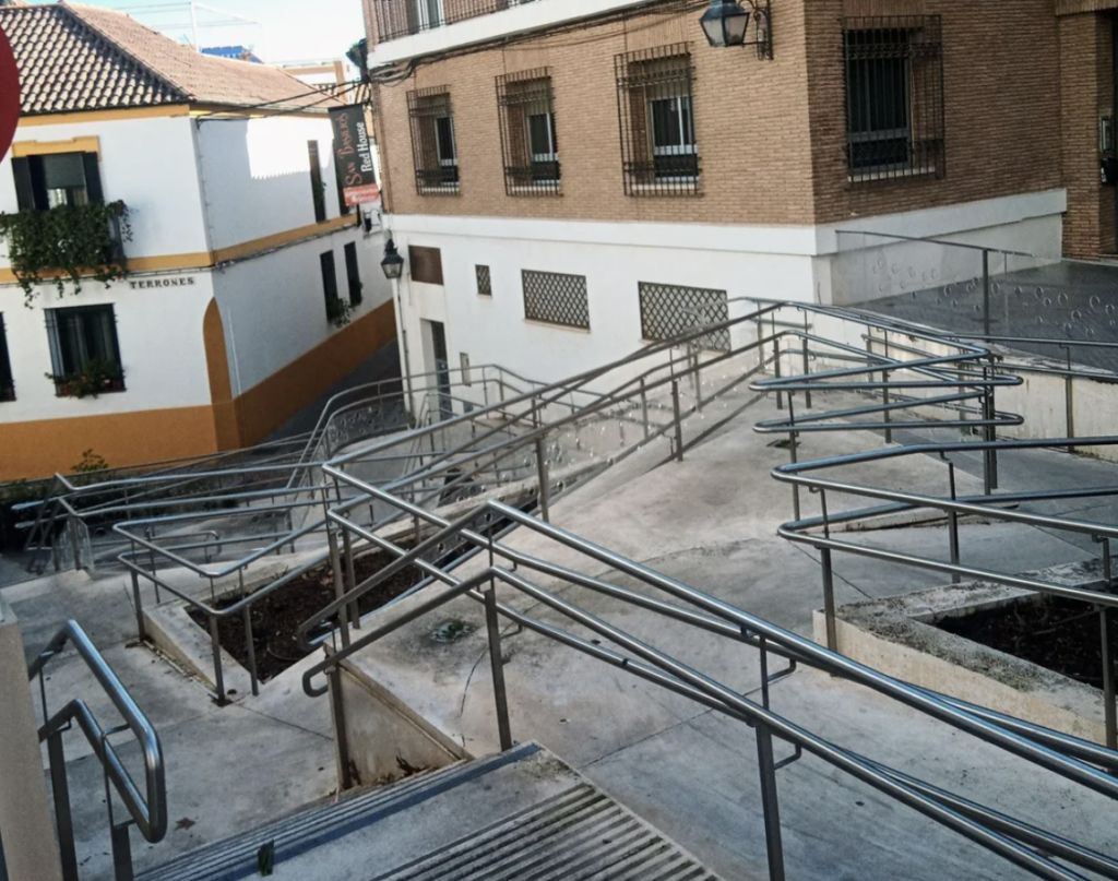 A complex network of zigzagging ramps with metal railings leads down to a lower street level between two buildings. The architecture is a mix of wooden and brick facades, with a small street and distant rooftops visible.