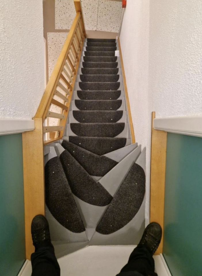 A staircase with narrow, irregularly-shaped steps, each covered in black carpet and featuring overlapping triangular designs in gray. The view is from the top, showing a wooden banister on the left and white walls. Two black shoes are visible at the bottom edge.