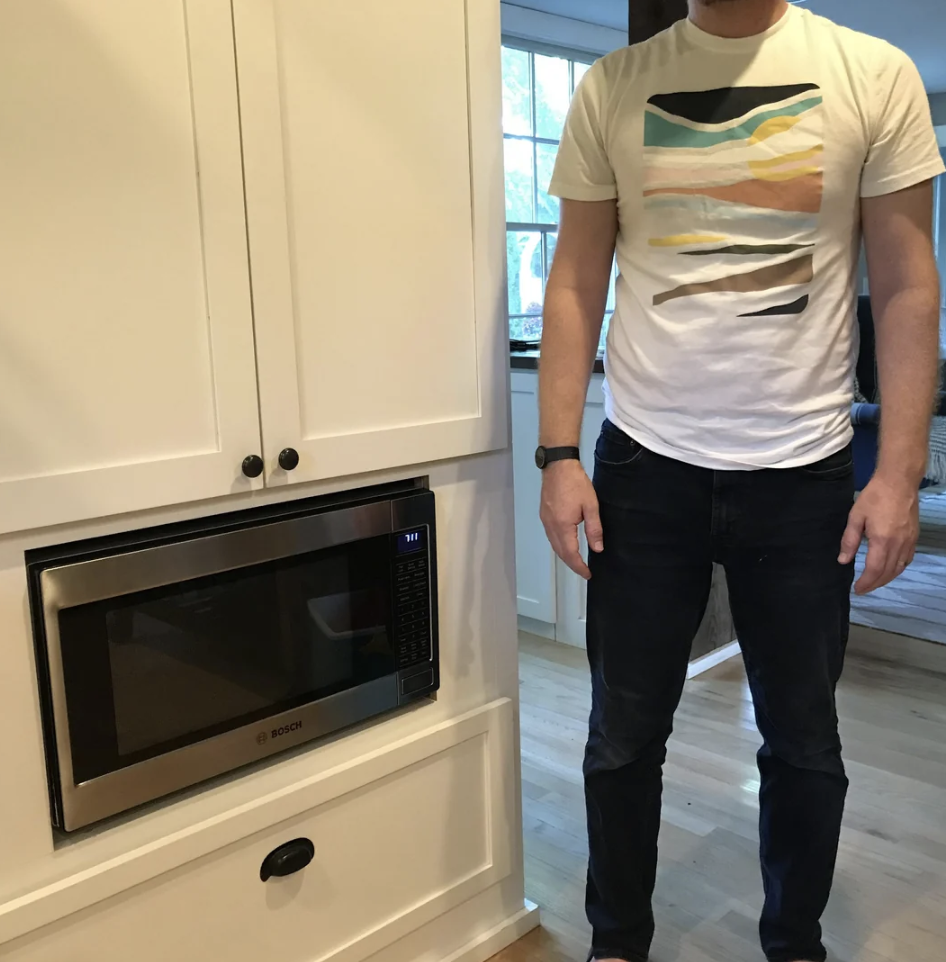 A person stands next to a built-in stainless steel Bosch microwave in a white cabinet. They wear a white t-shirt with abstract pastel shapes and dark jeans. The room has wooden floors and a window in the background.