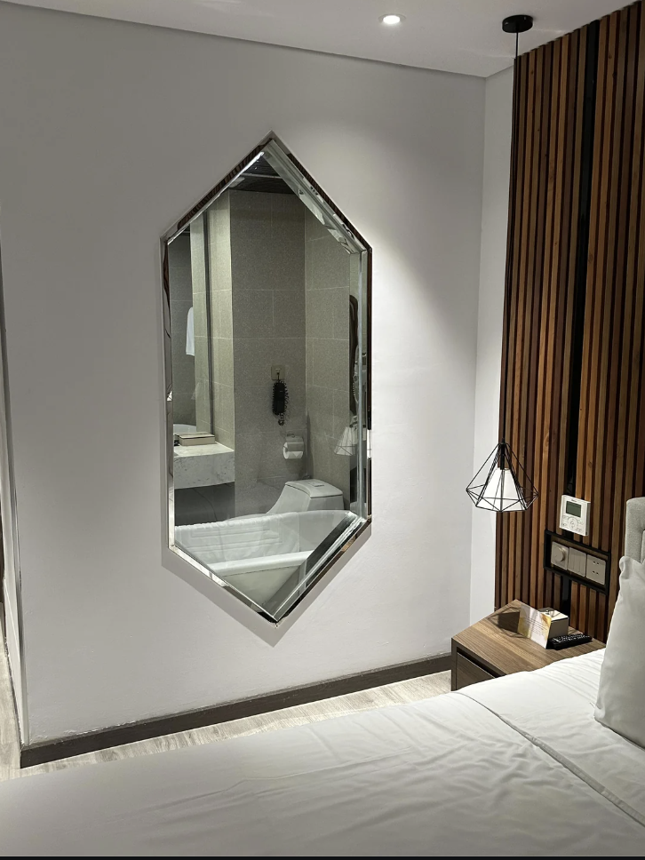 Modern hotel room with a geometric mirror reflecting a bathroom. The wall features vertical wooden slats, and there's a hanging lamp above a small bedside table. The bed is partially visible with white linens.