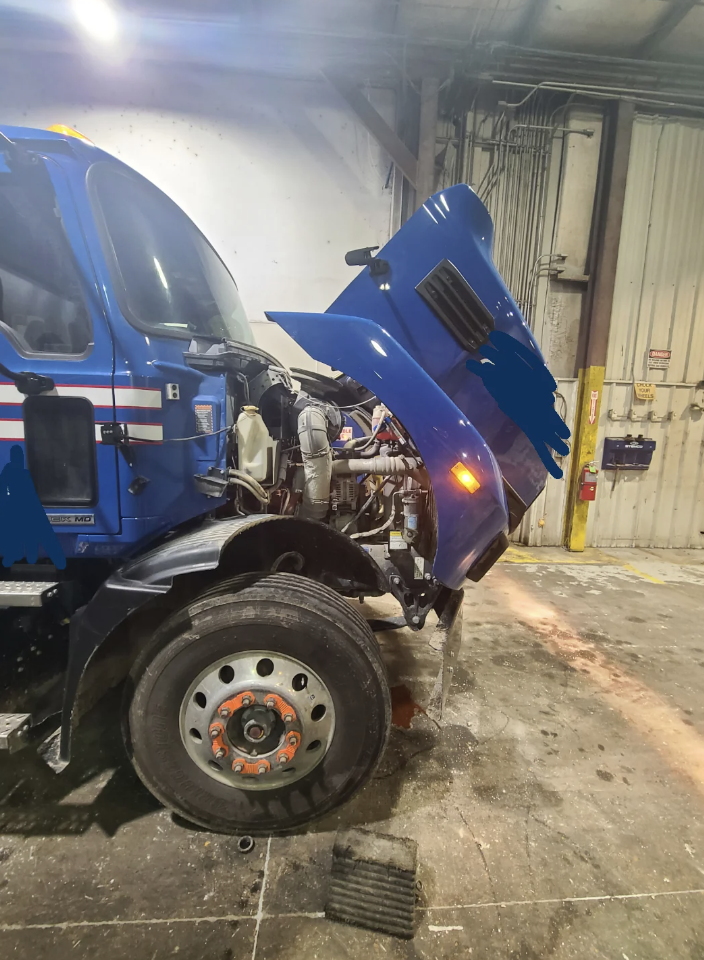 A blue truck with its hood open is parked inside a garage. The engine is visible, and tools are scattered on the floor. The garage has a white wall and metal door in the background.