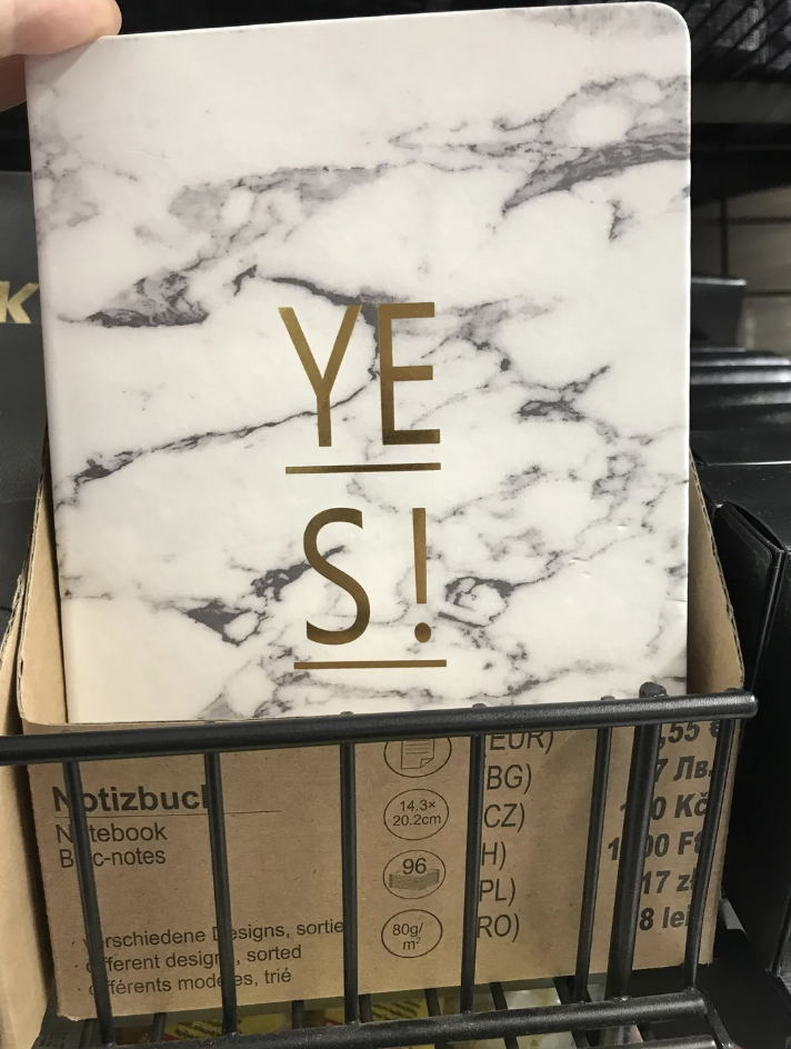 A marble-patterned notebook with the word "YES!" in gold letters is displayed upright in a shelf. Behind it, packaging shows dimensions and specifications in multiple languages.