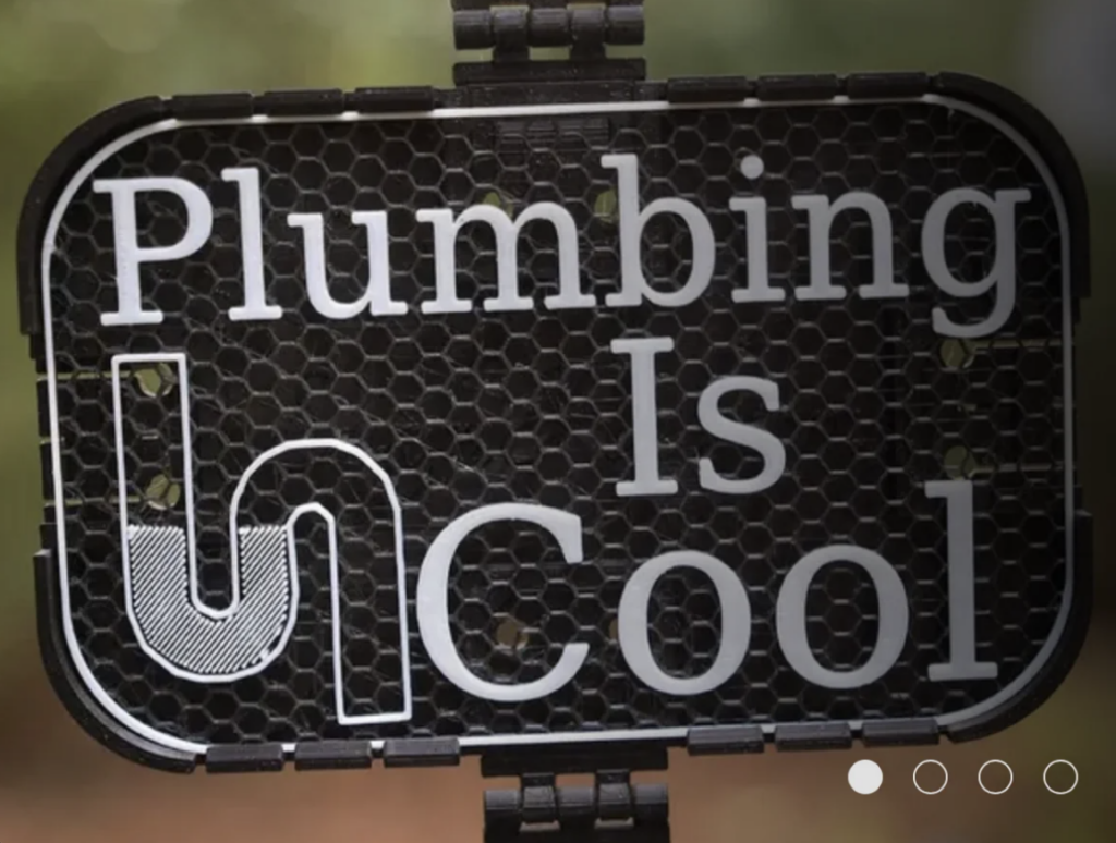 A dark sign with a honeycomb pattern, displaying the text "Plumbing Is Cool" in bold white letters. The letter "P" is styled as a pipe. The background is blurred with hints of greenery.